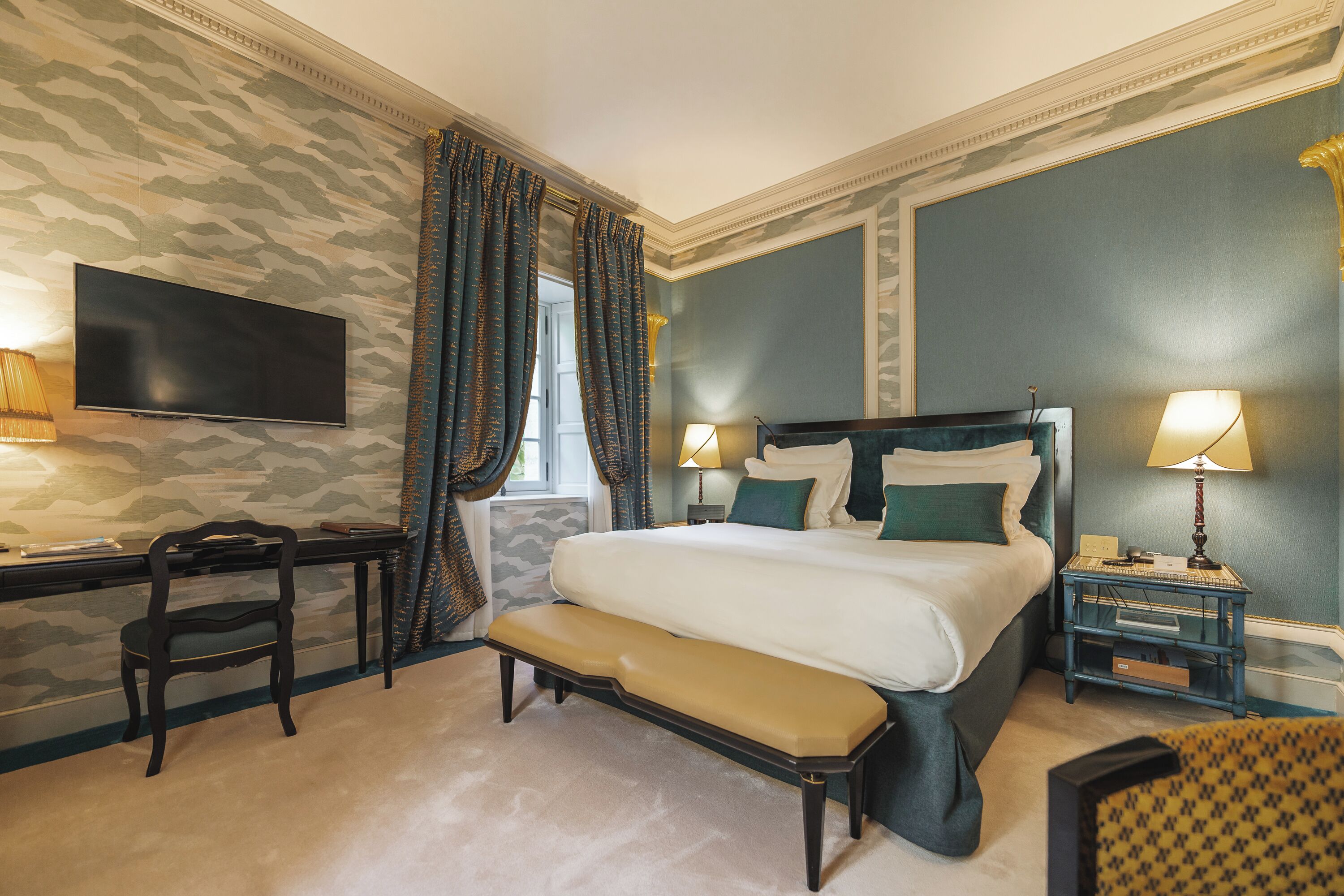A hotel room with a bed, a desk, a chair, a bench, and two bedside tables with lamps.