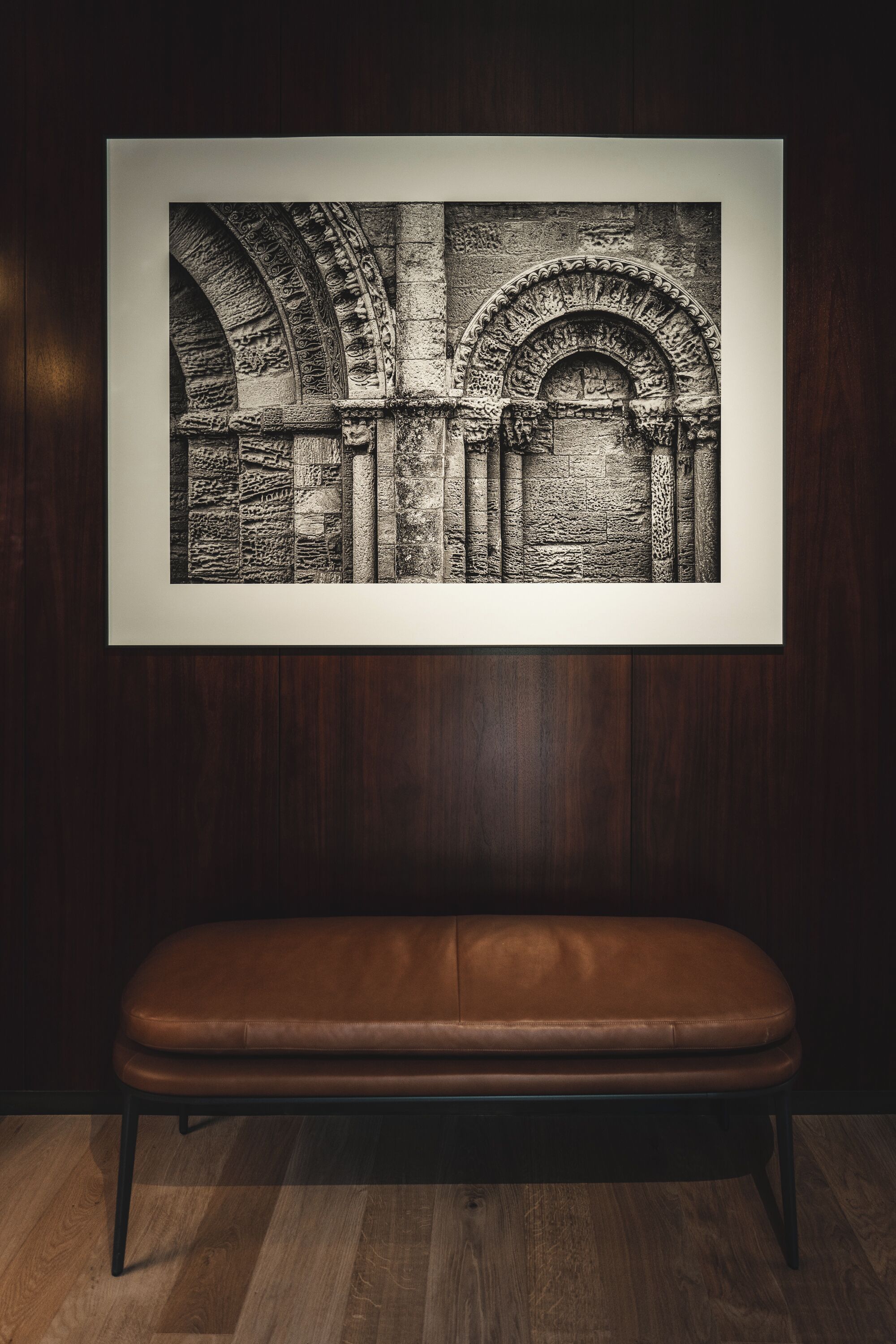 A framed black and white photograph of an arched stone structure on a wall, with a brown leather bench in front.