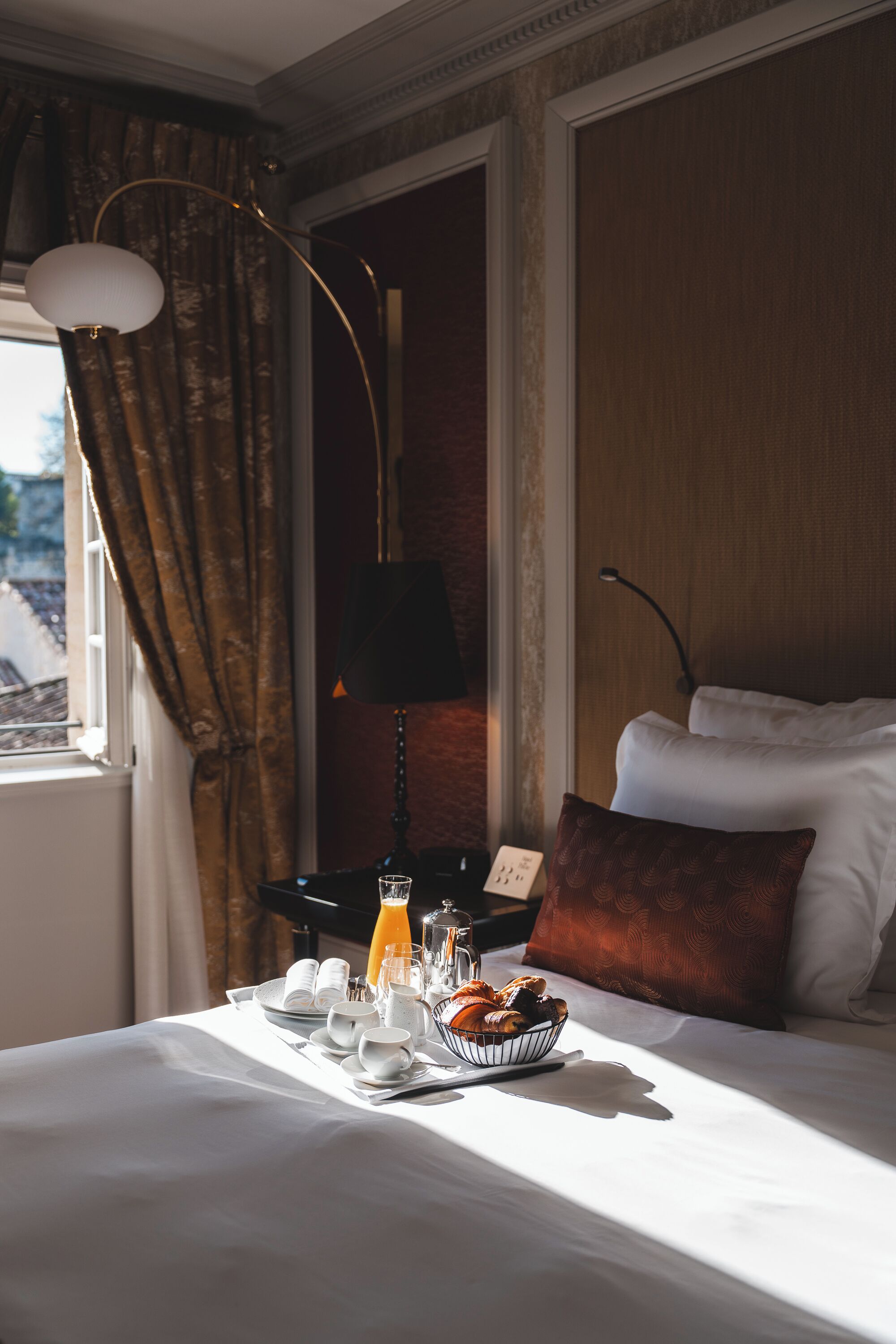 A neatly made bed with white linens, a brown pillow, and a bedside table with a lamp, a glass of orange juice, and a bowl of pastries.