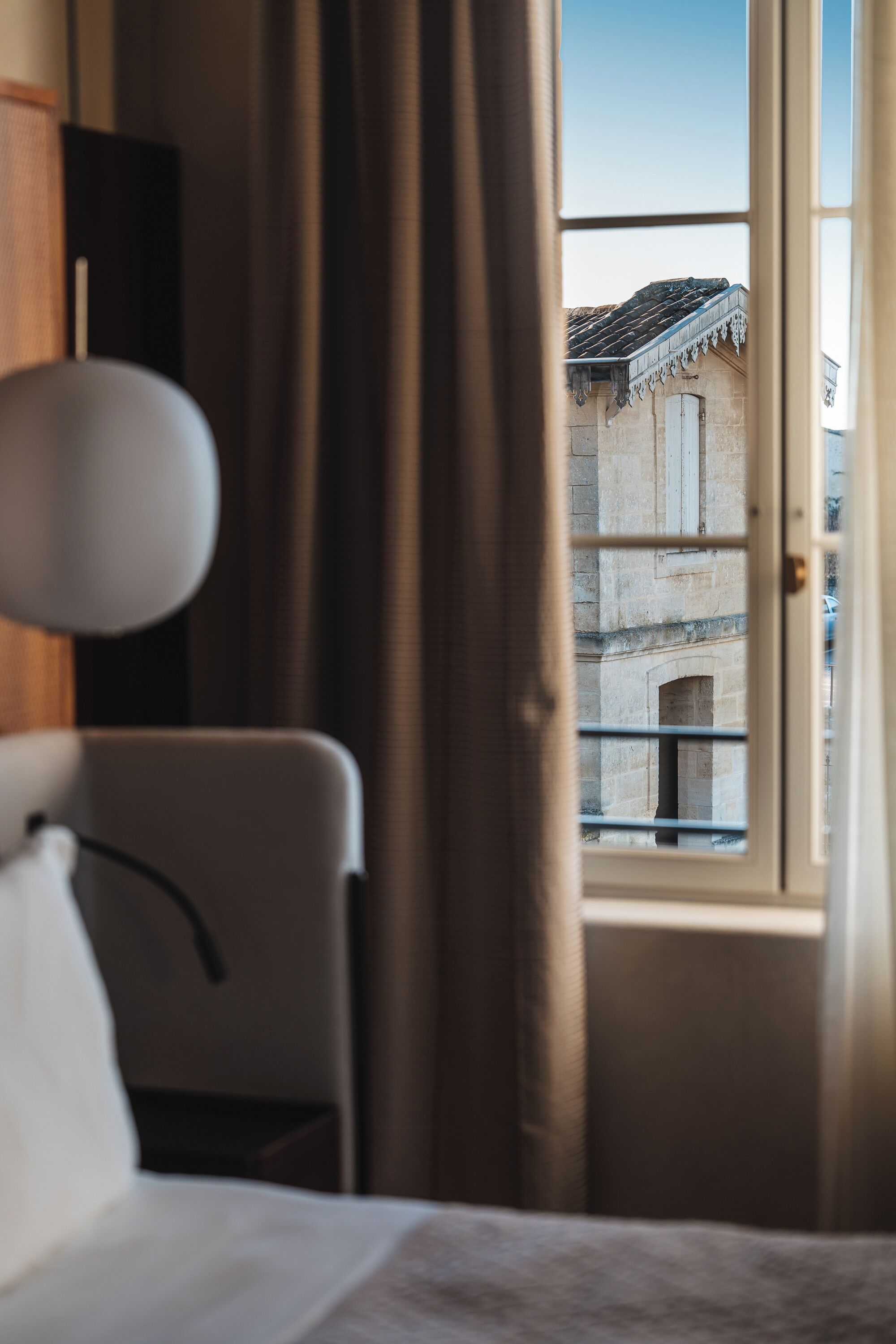 Quarto de hotel com cama, cadeira, luminária e janela com cortinas.