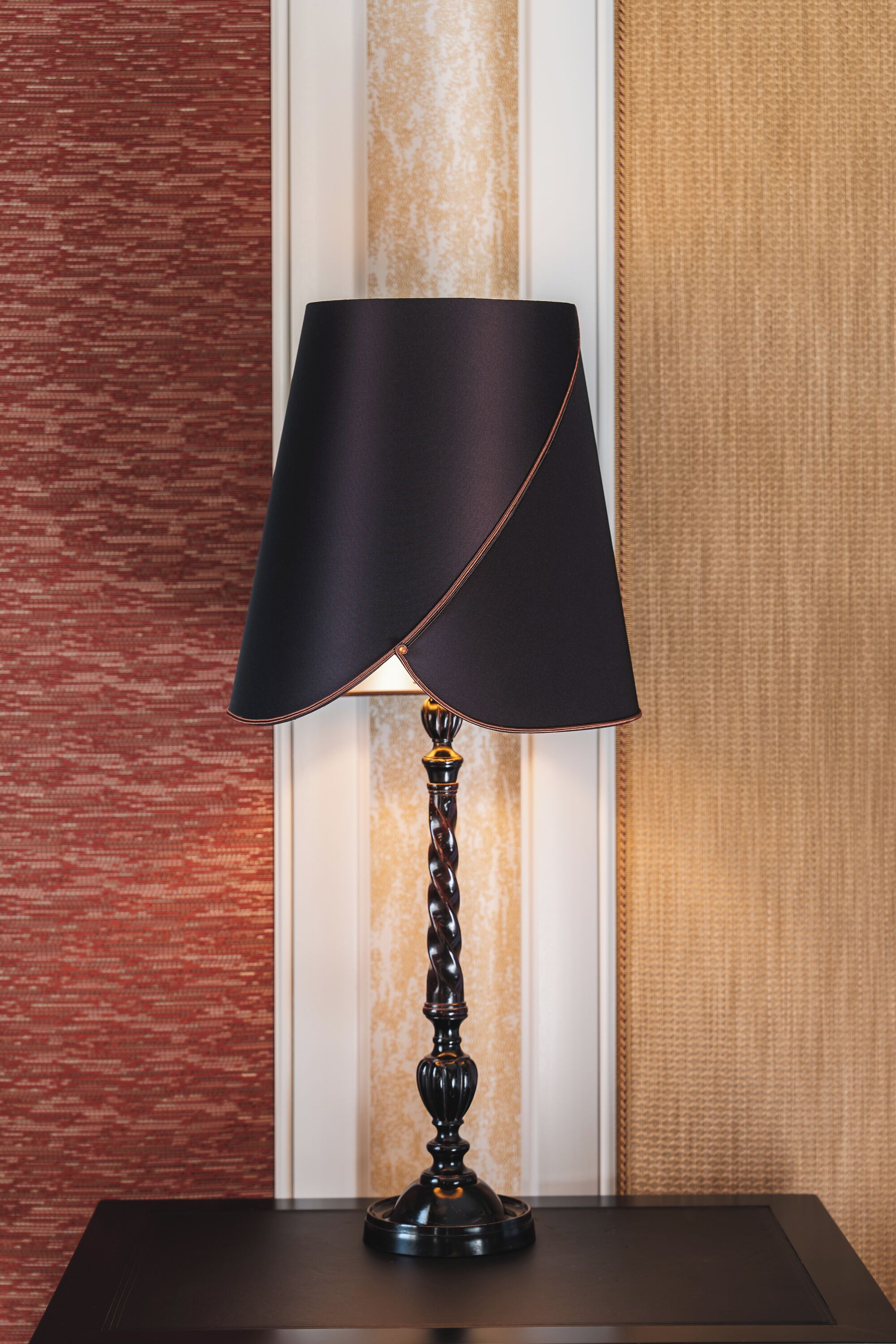 Una lámpara de mesa con pantalla oscura sobre una mesa de madera frente a una pared con textura.