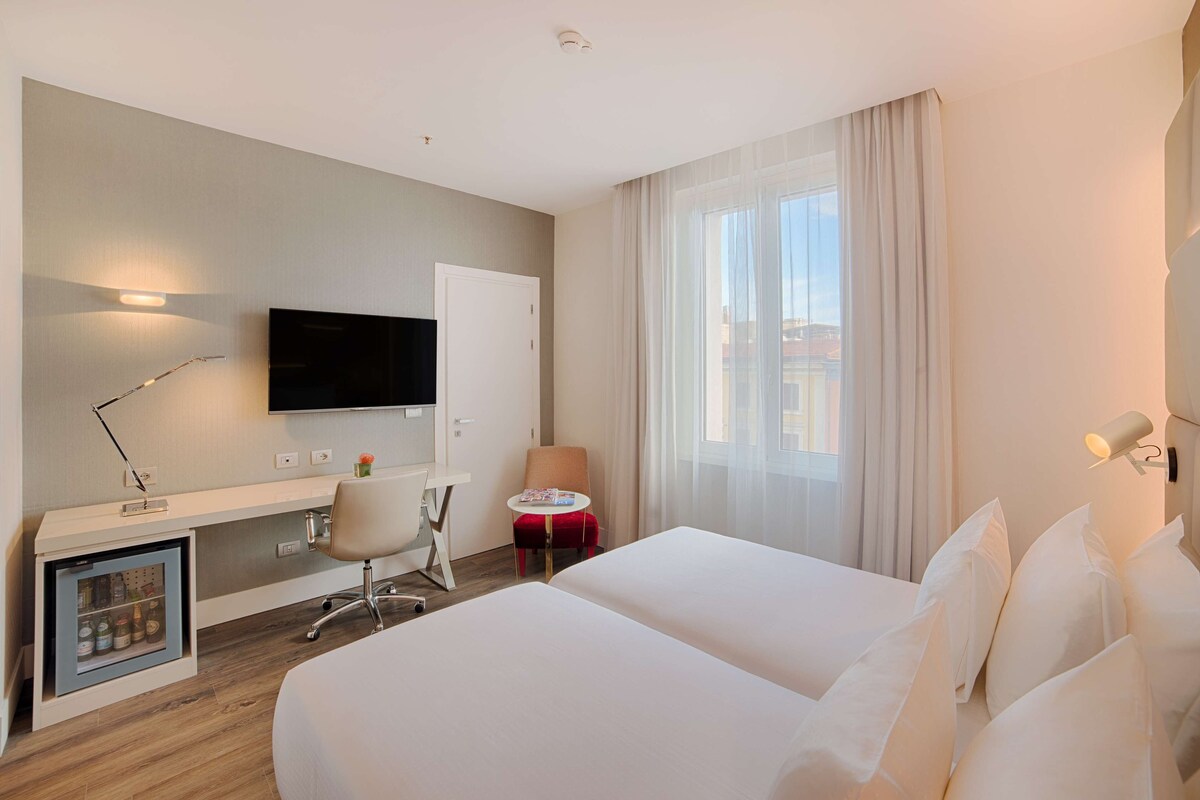 A hotel room with a bed, a desk, a chair, a TV, and a window with curtains.