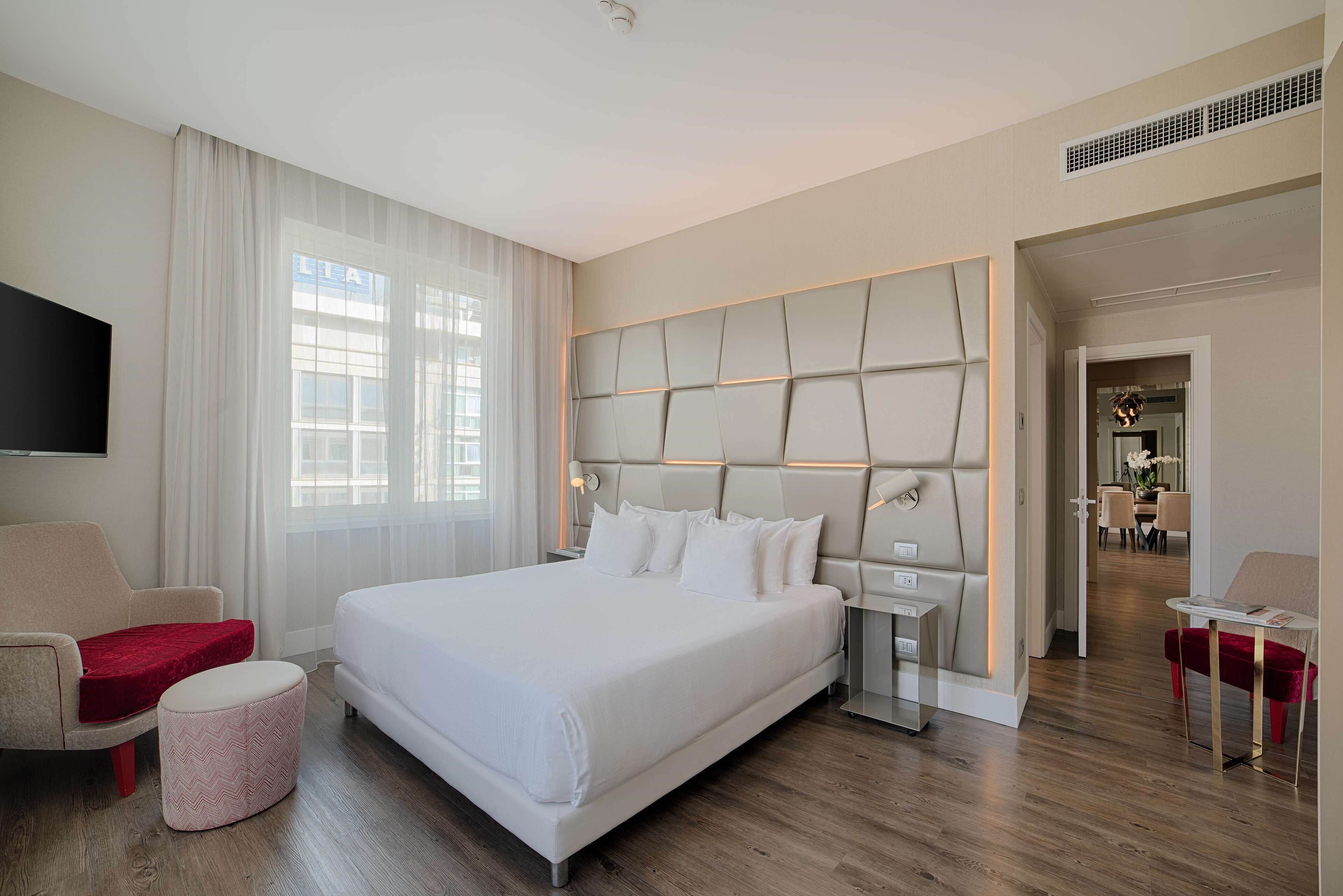 A modern hotel room with a large bed, a TV, a seating area, and a view of the city.