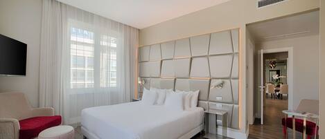 A modern hotel room with a large bed, a TV, a seating area, and a view of the city.