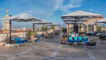 Un área de estar al aire libre con varios paraguas, sofás y sillones, dispuesta en un patio con baldosas y vistas a la ciudad.