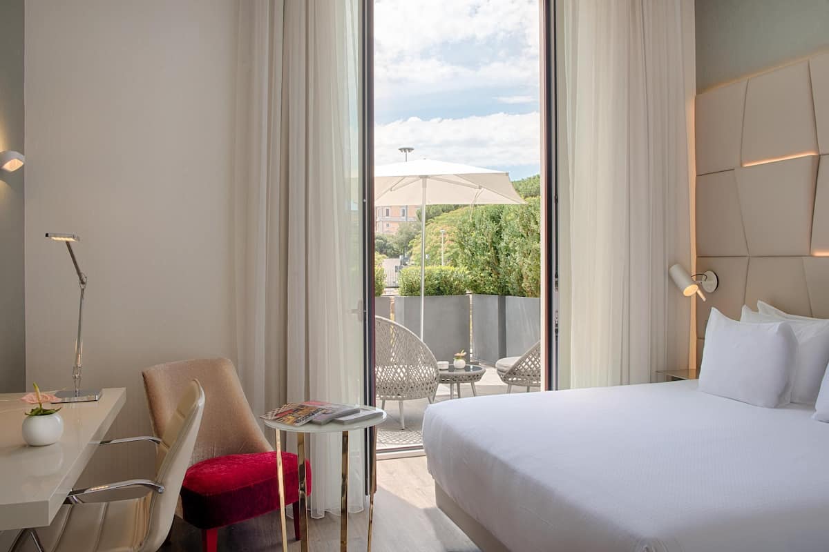 A hotel room with a bed, a desk, a chair, a small table with a magazine, and a view of the outside.