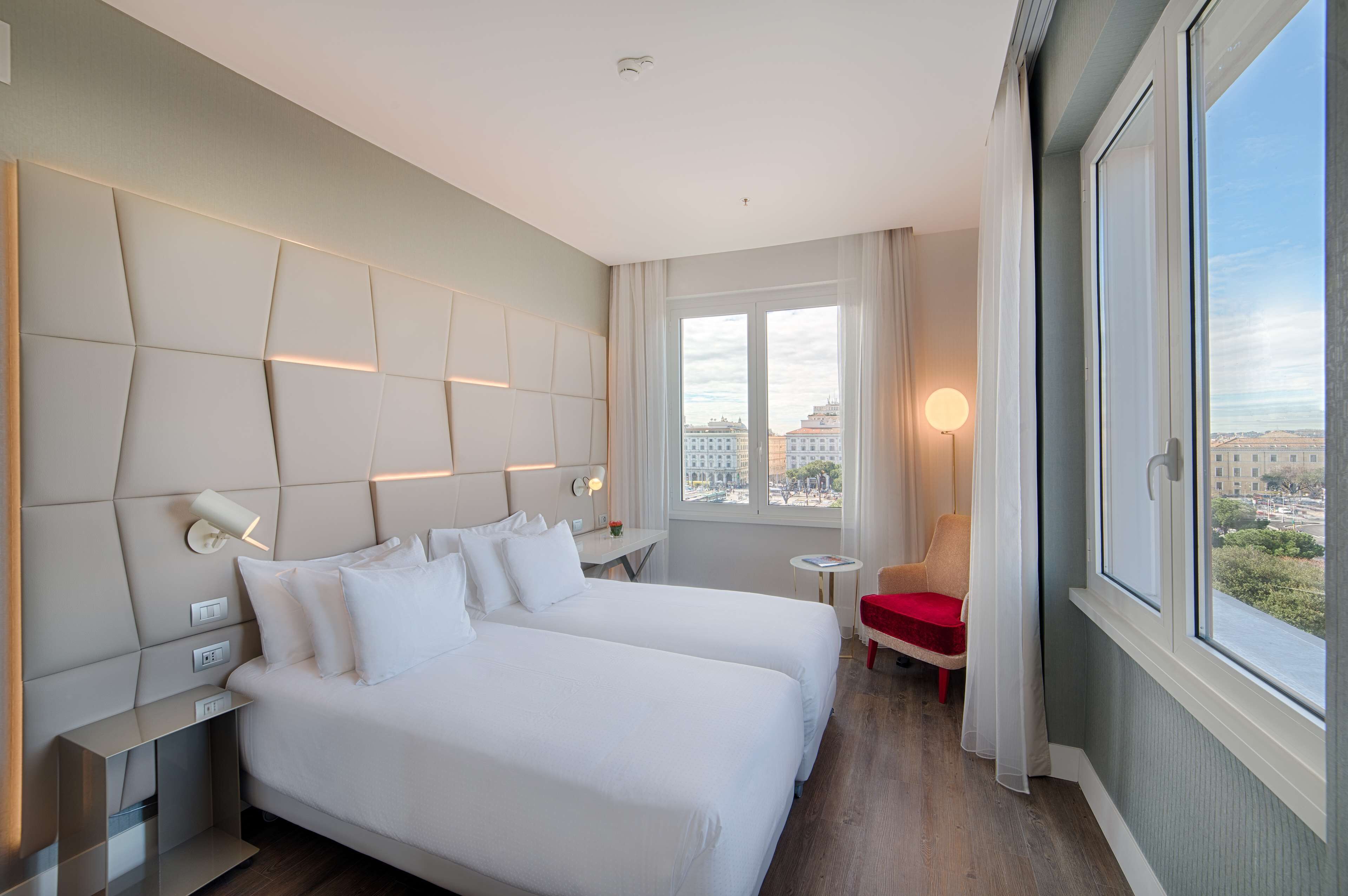 Ein Hotelzimmer mit zwei Betten, einem kleinen Tisch, einem Sessel und Blick auf die Stadt durch große Fenster.