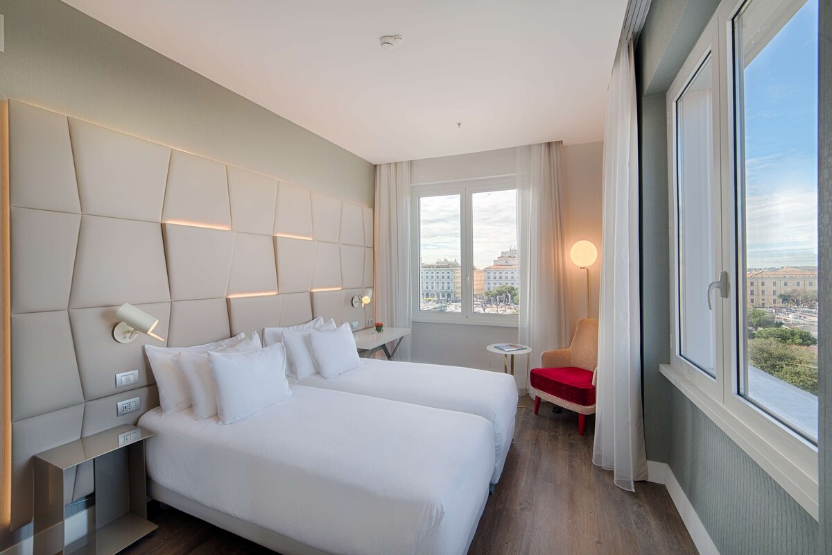A hotel room with two beds, a small table, a chair, and a view of the city through large windows.