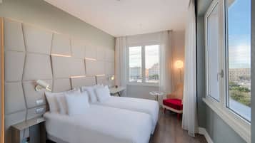 Ein Hotelzimmer mit zwei Betten, einem kleinen Tisch, einem Sessel und Blick auf die Stadt durch große Fenster.