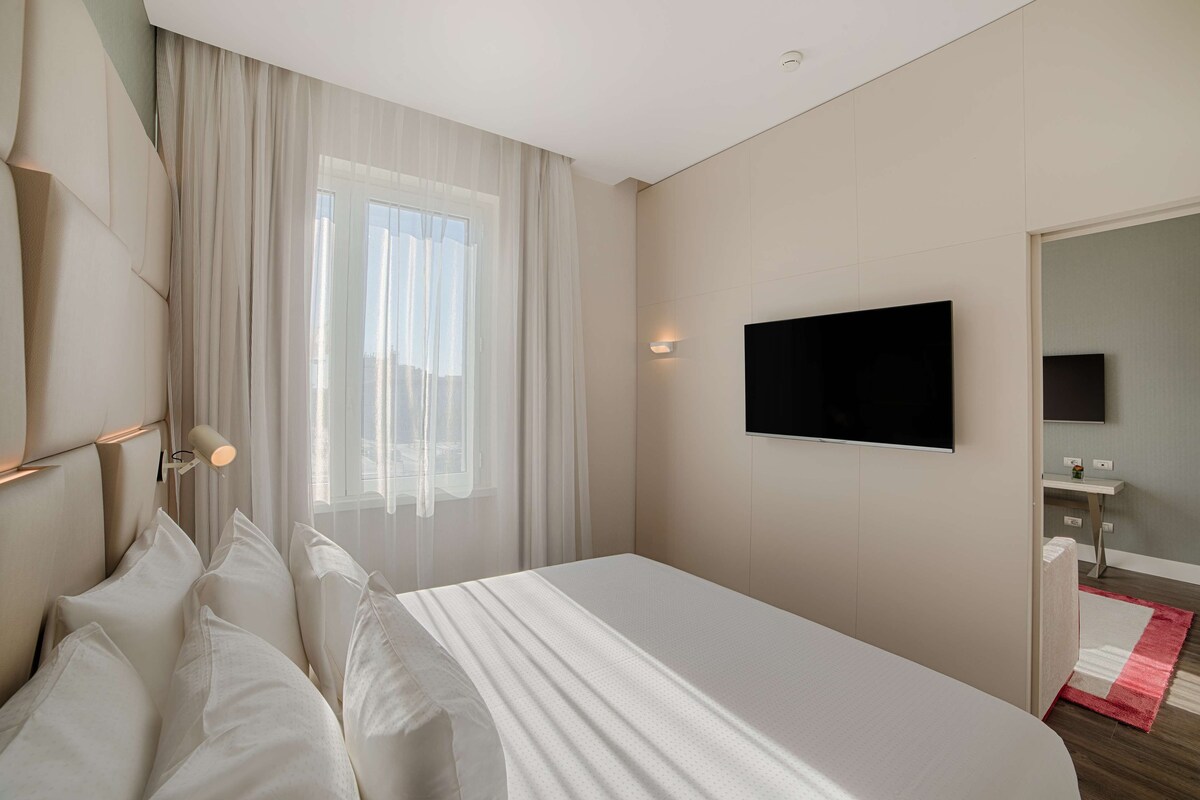 A modern hotel room with a large bed, a flat-screen TV mounted on the wall, a bedside table, and a window with sheer curtains.