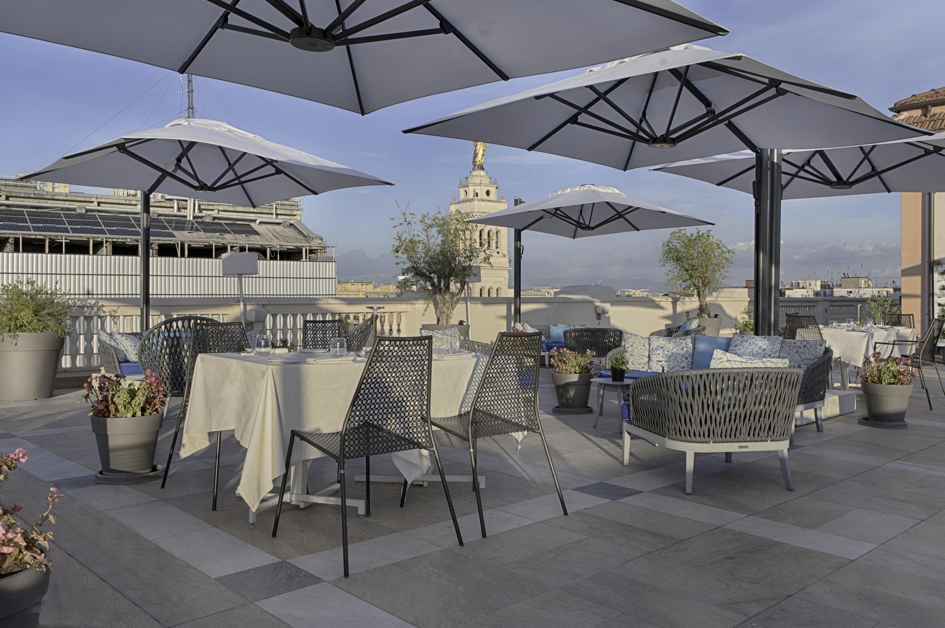 A rooftop terrace with tables and chairs under umbrellas, potted plants, and a view of a historic building with a tower.