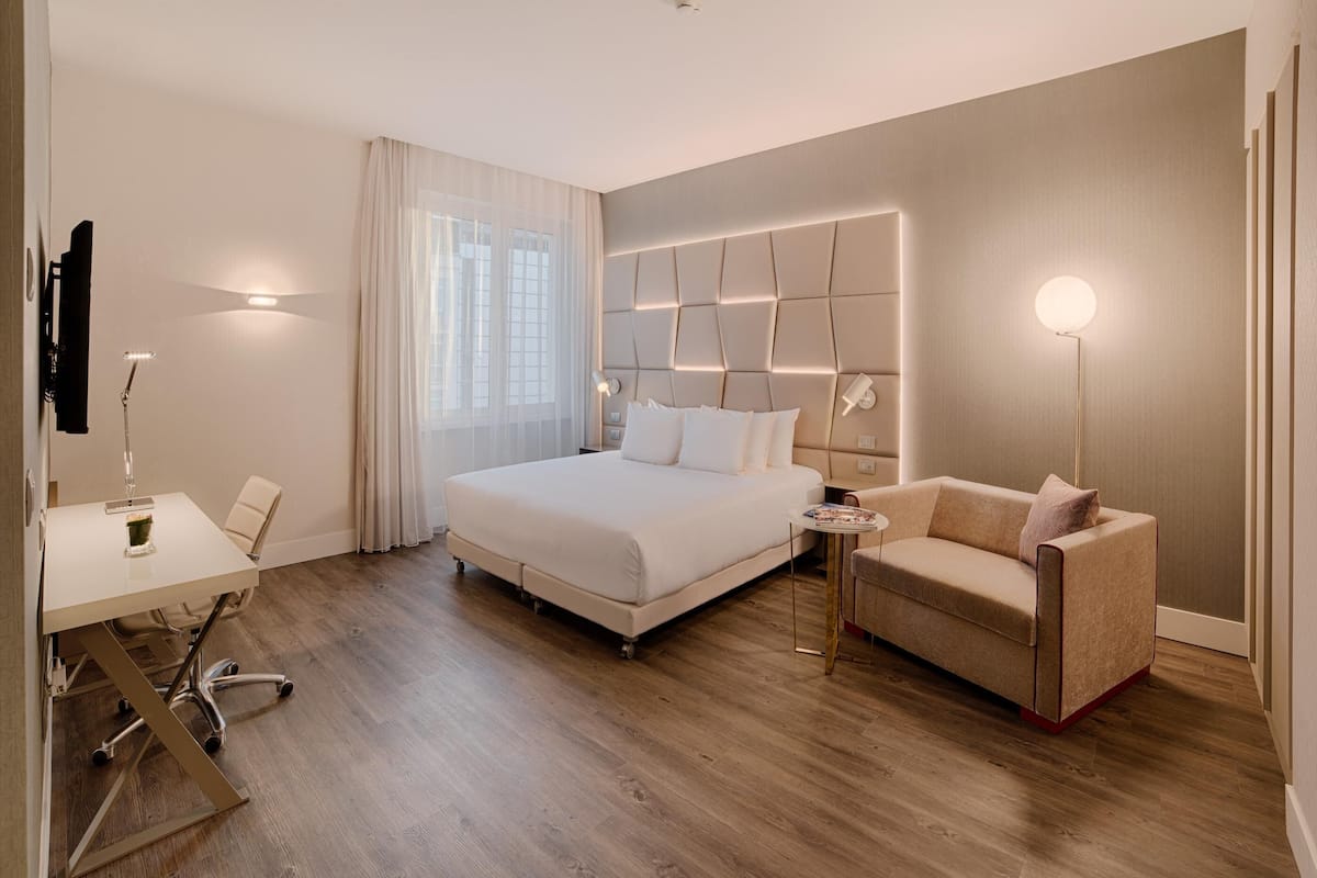 A modern hotel room with a large bed, a desk, a sofa, and a bedside table.