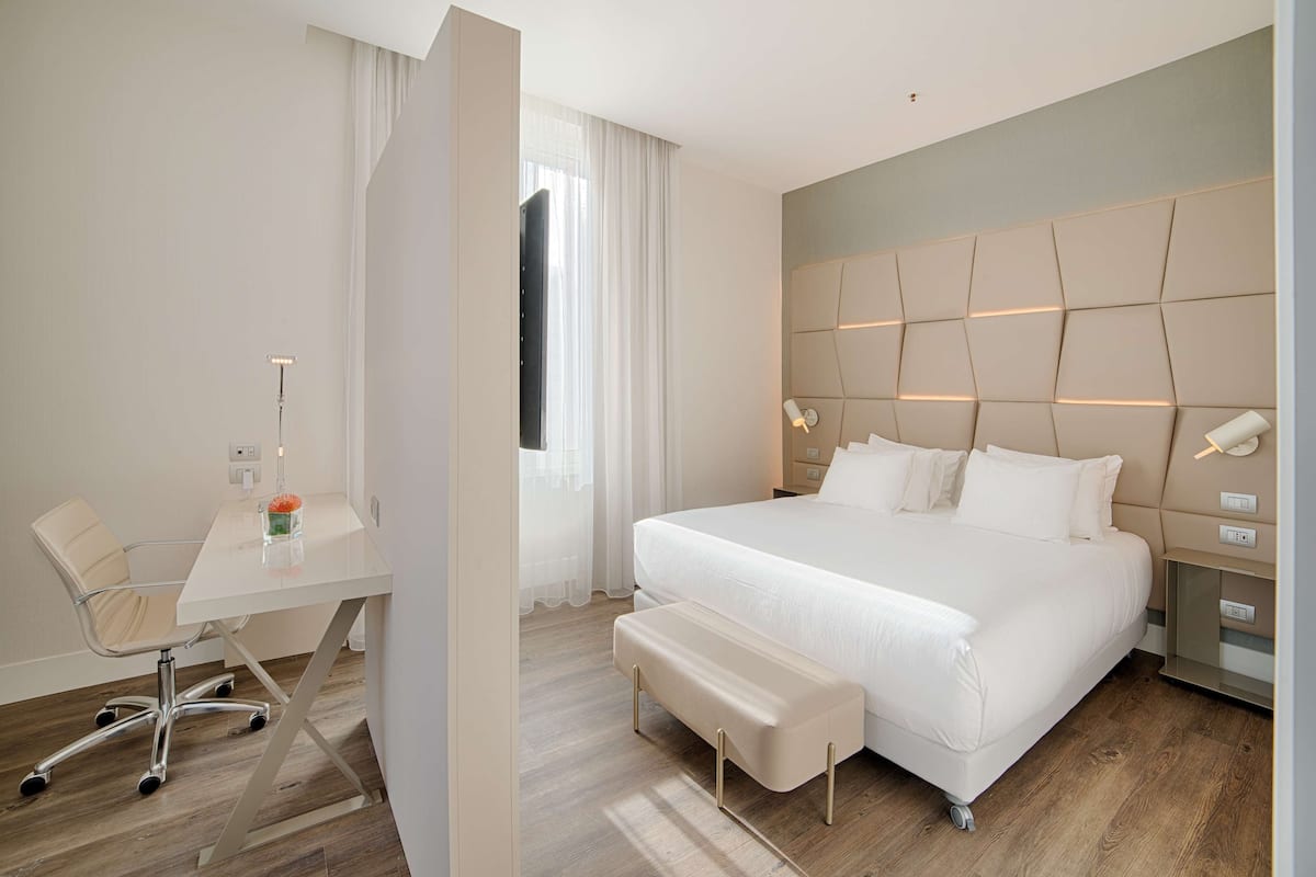 A modern hotel room with a large bed, a desk with a chair, and a bench.