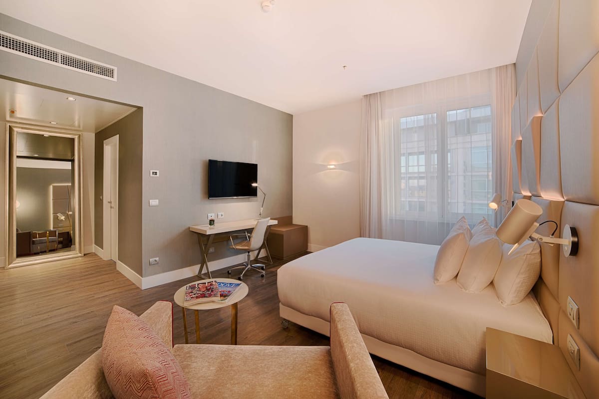 A modern hotel room with a large bed, a desk with a chair, a television, and a window with curtains.