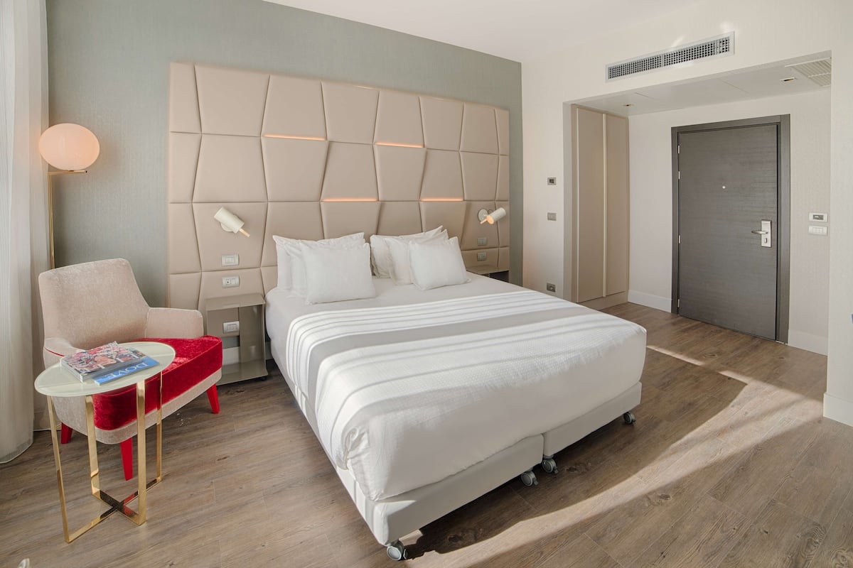 A modern hotel room with a large bed, a red armchair, a small table, and a bedside lamp.