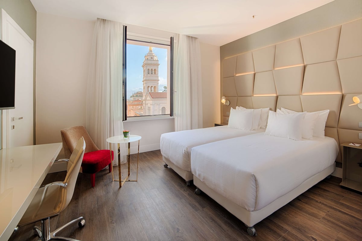 A hotel room with two beds, a desk, a chair, a small table, and a window with a view of a building.