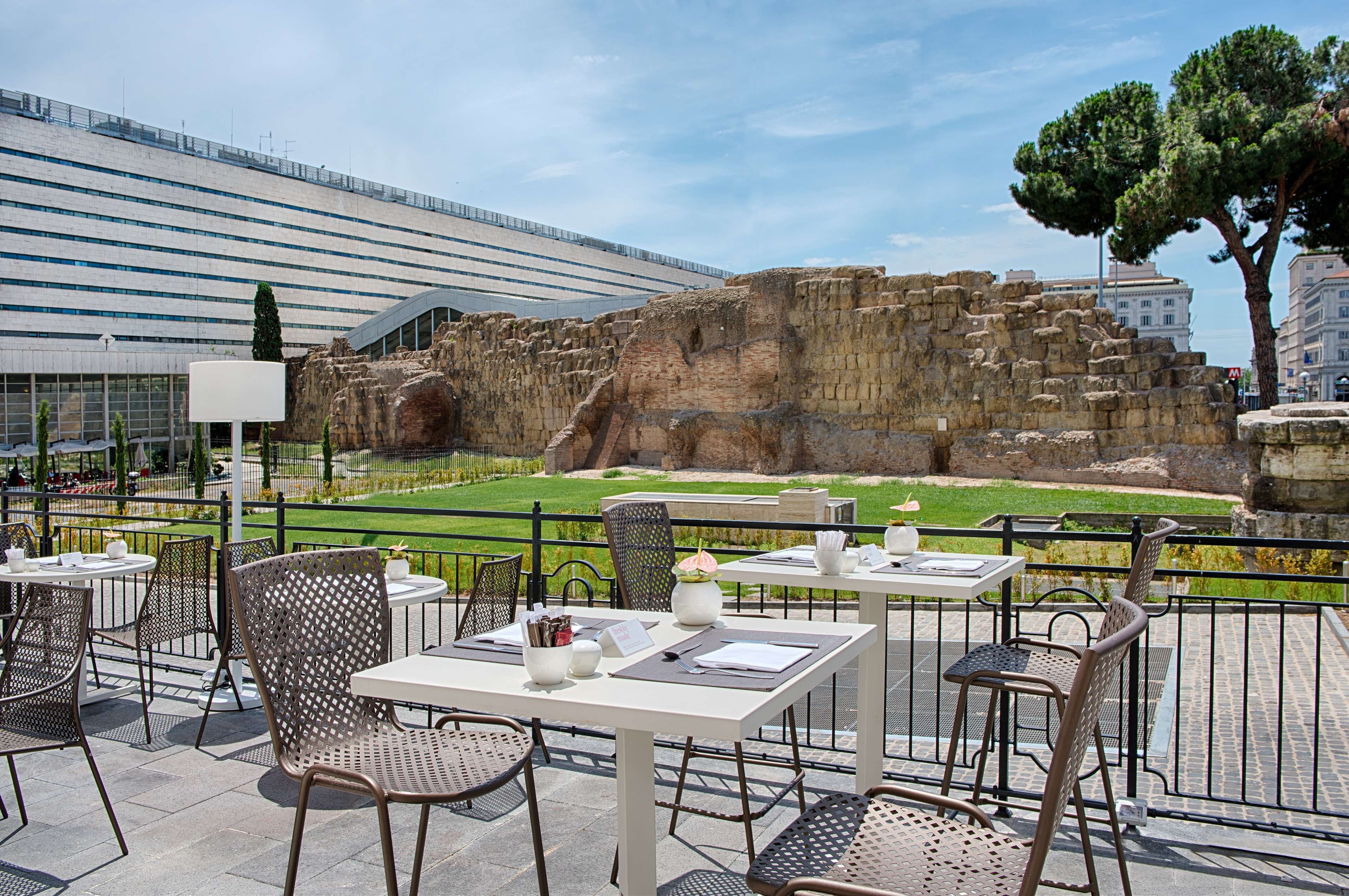 An outdoor cafe with tables and chairs, overlooking ancient ruins and a modern building.