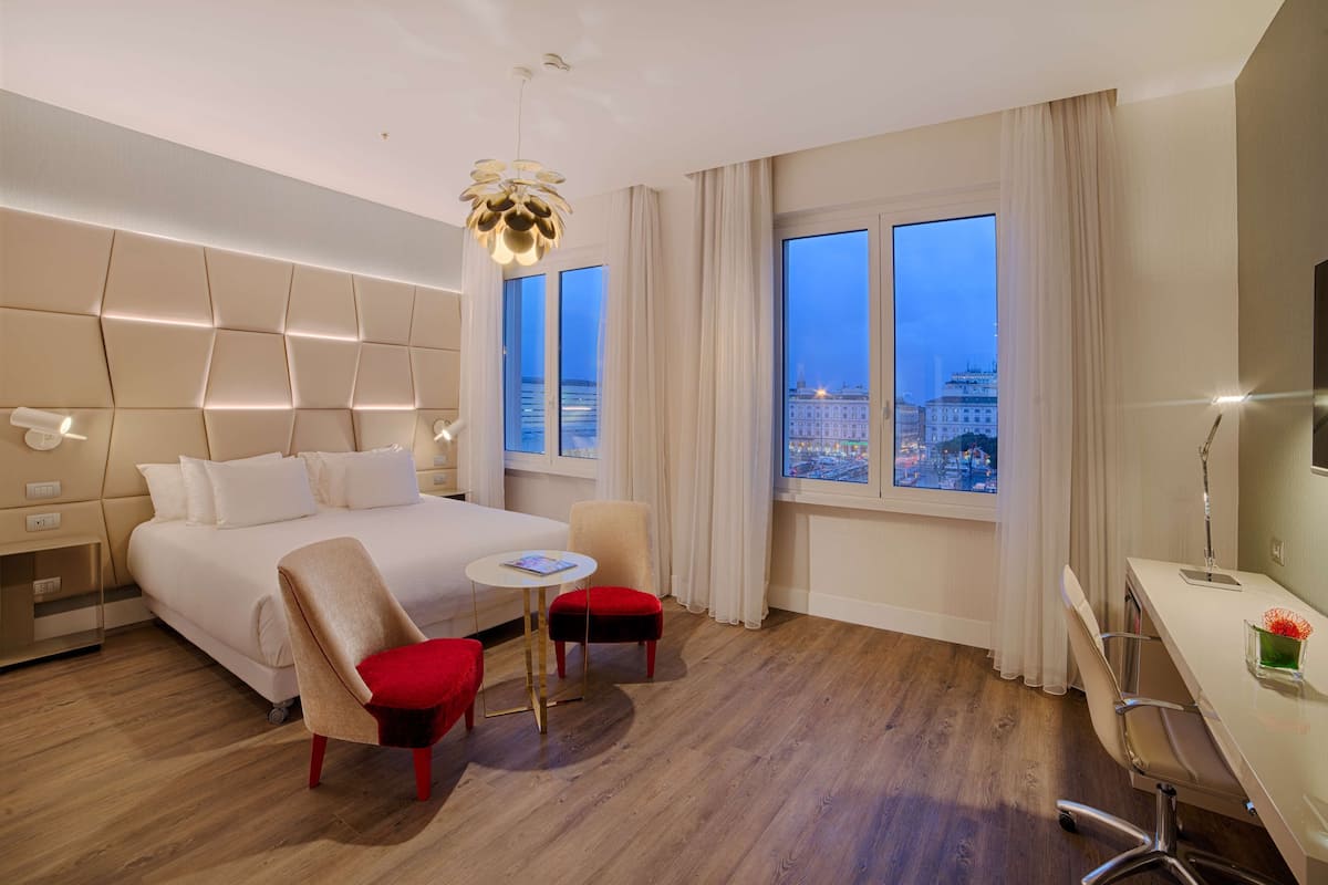 A modern hotel room with a large bed, a desk, a chair, and a view of a cityscape.