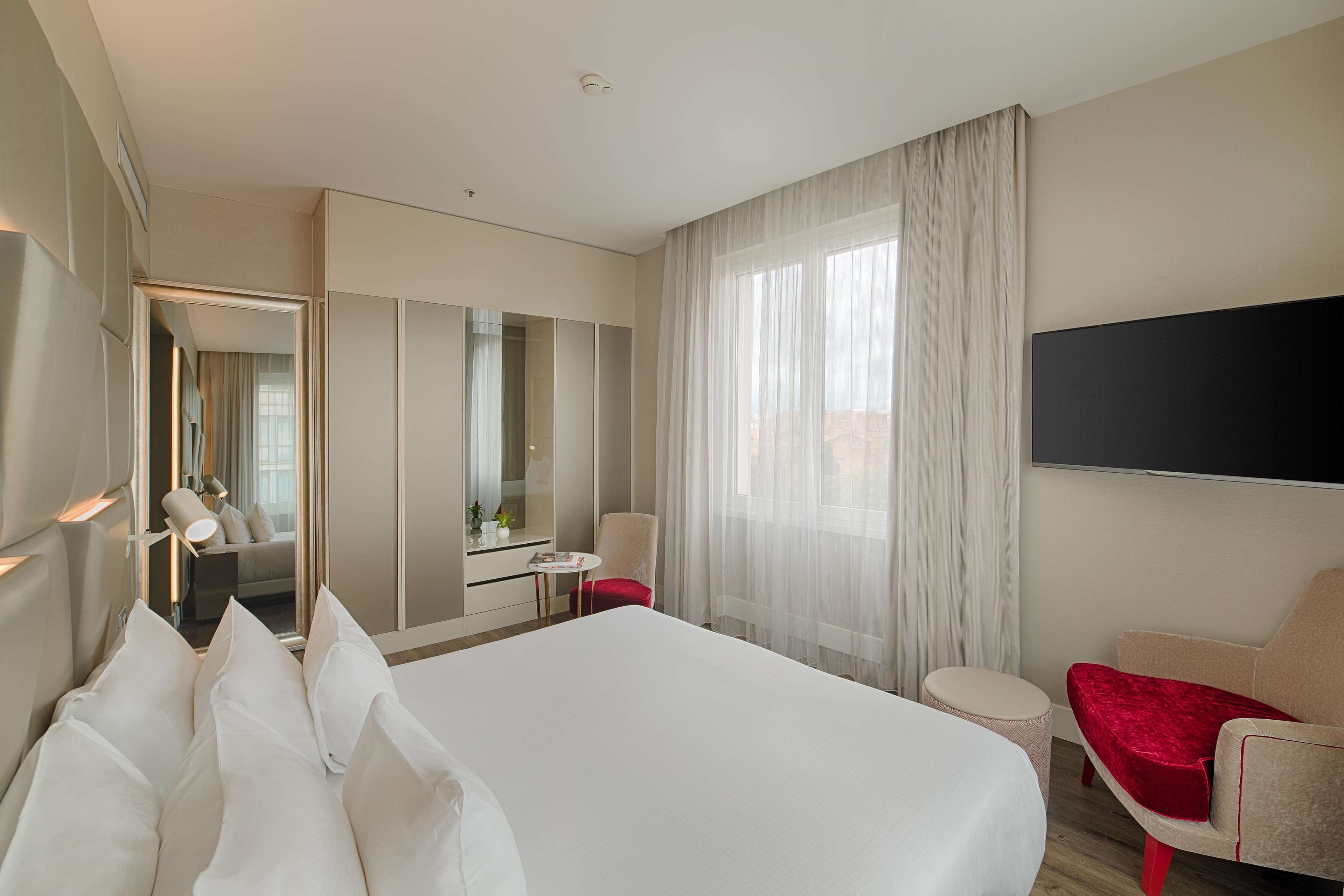 A hotel room with a large bed, a desk, a chair, a TV, and a view of the city.