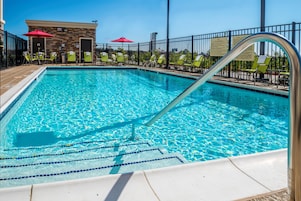 A swimming pool with a diving board, surrounded by lounge chairs and a building with a brick facade.