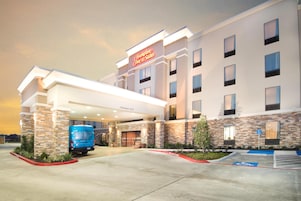 Un hotel Hampton Inn & Suites con entrada cubierta, una furgoneta de reparto azul y cielo despejado.
