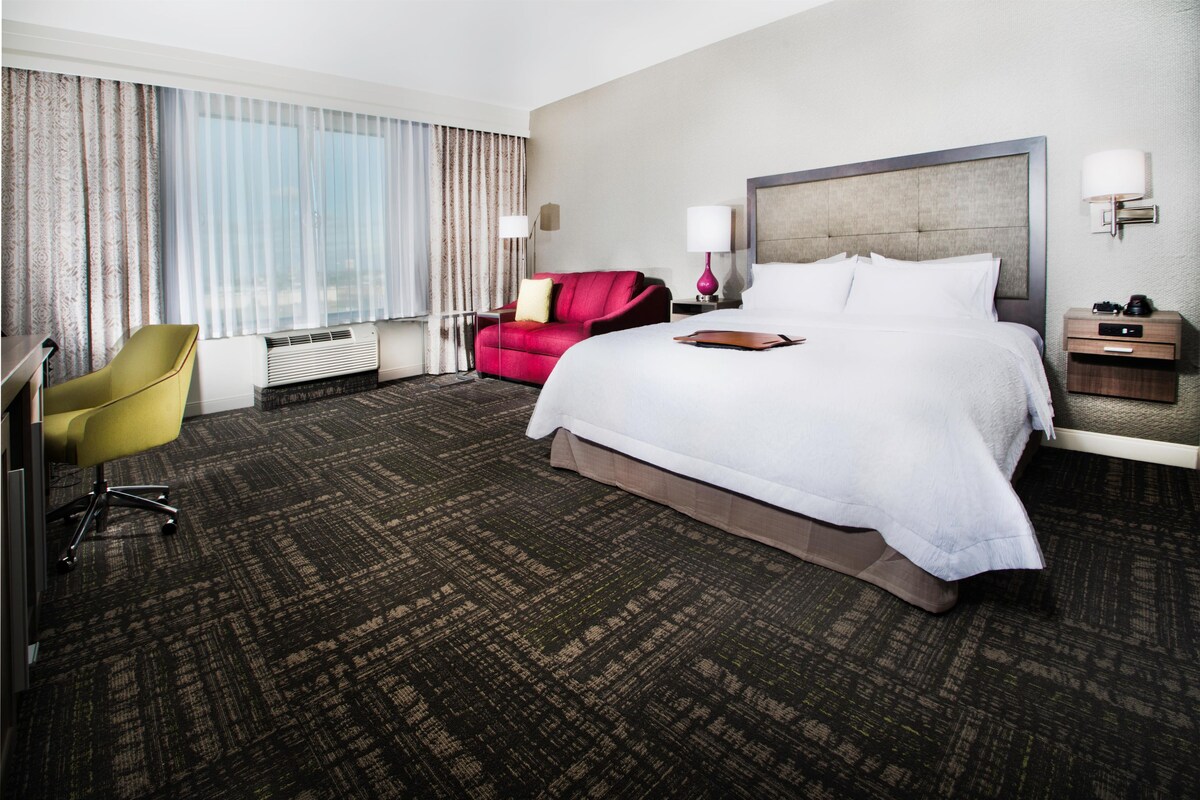 A hotel room with a large bed, a desk, a chair, and a window with curtains.