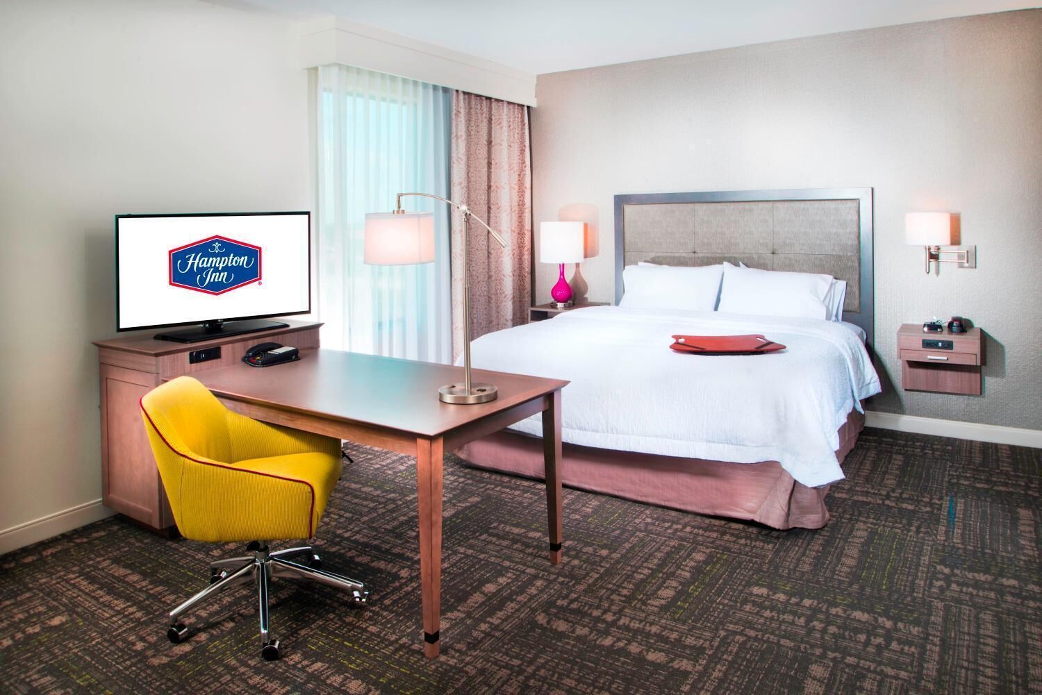 A hotel room with a bed, a desk with a chair, a television, and a nightstand with lamps.
