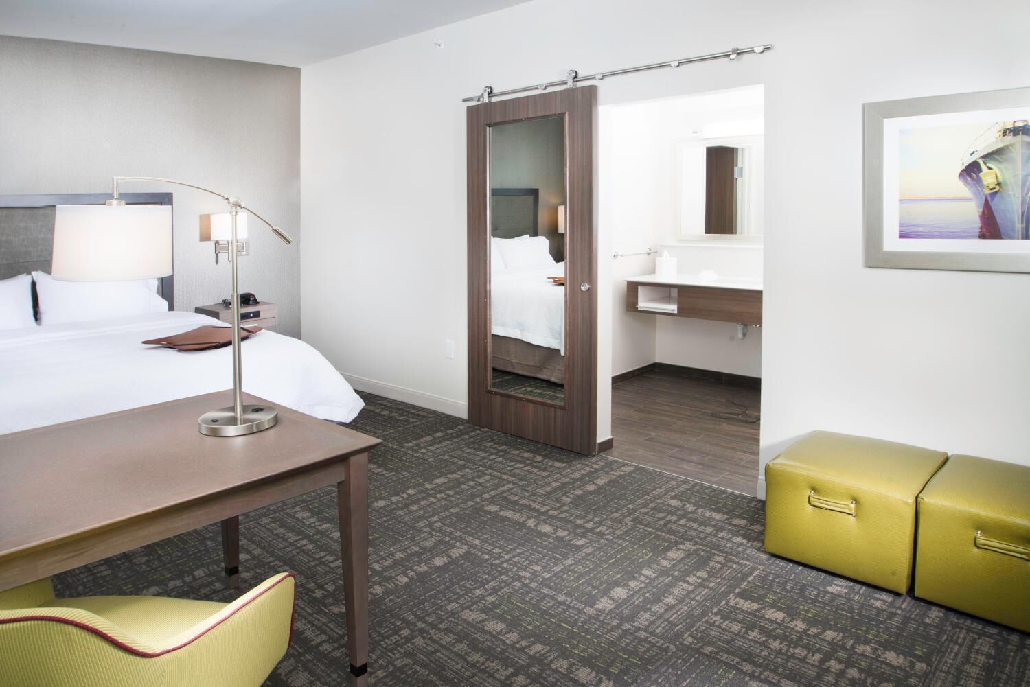 A hotel room with a bed, bedside table, lamp, chair, wardrobe, and a bathroom with a sink and mirror.