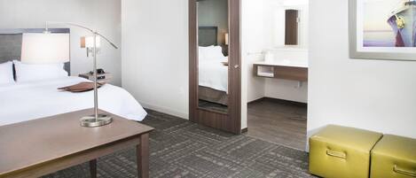 A hotel room with a bed, bedside table, lamp, chair, wardrobe, and a bathroom with a sink and mirror.