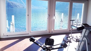 A gym with exercise equipment and large windows overlooking a body of water. - Hotel Garni Battello (Melide)