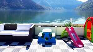 A terrace with a sofa, children's play equipment, and a view of a lake with mountains in the background. - Hotel Garni Battello (Melide)