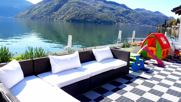 A checkered outdoor seating area with a sofa and cushions, a children's play structure, and a view of a lake with mountains in the background. - Hotel Garni Battello (Melide)