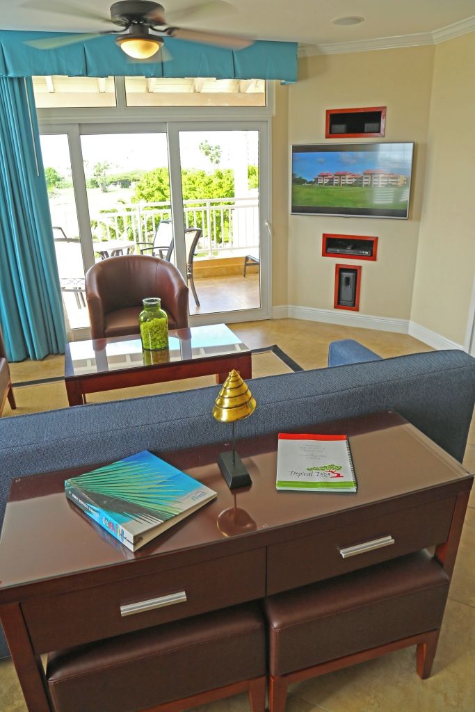 A room with a sofa, a table with a book and a lamp, a TV on the wall, and a view of the outdoors through a sliding door.