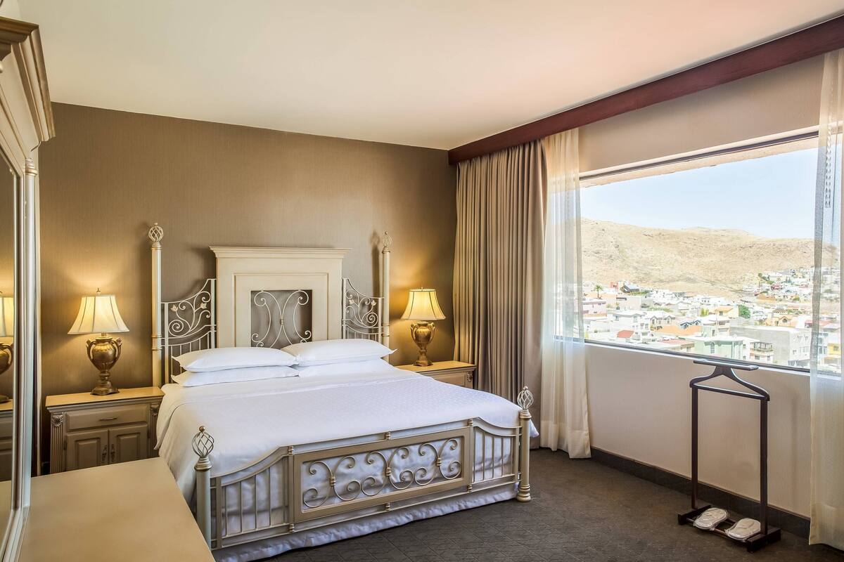 A hotel room with a large bed, two bedside tables with lamps, a dresser, and a view of a cityscape through a large window.