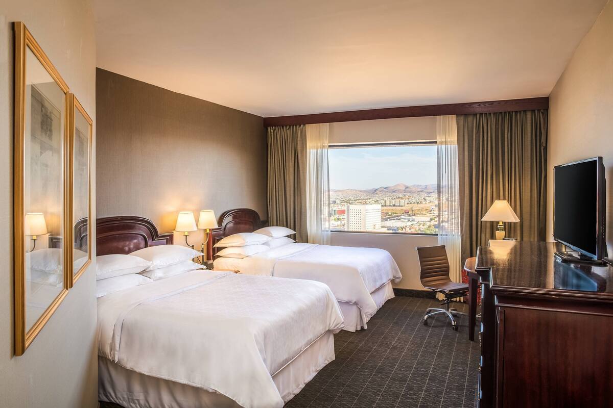 A hotel room with two beds, a desk, a TV, and a window with a city view.