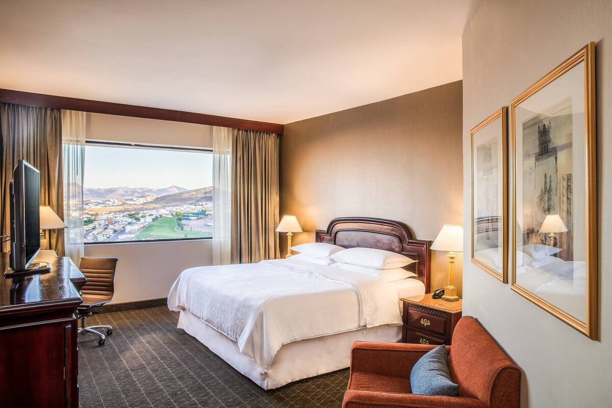 A hotel room with a large bed, two bedside tables with lamps, a desk with a chair, a TV, and a view of a cityscape through a large window.