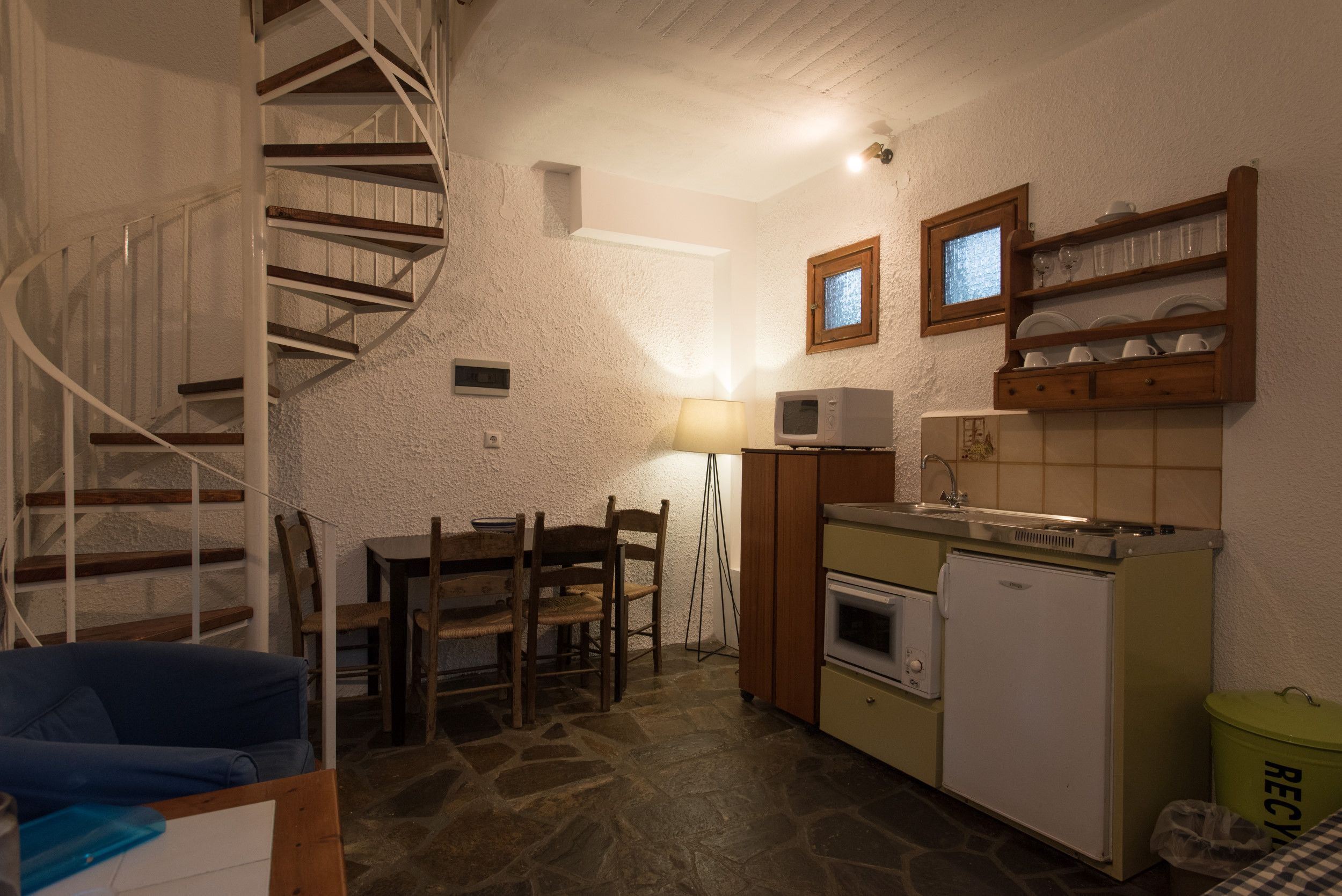A compact kitchen with a spiral staircase, a dining table with chairs, a microwave, a sink, and a small refrigerator.