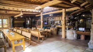 Un interior rústico de madera con largas mesas comunes y bancos, una zona de bar y una chimenea de piedra.