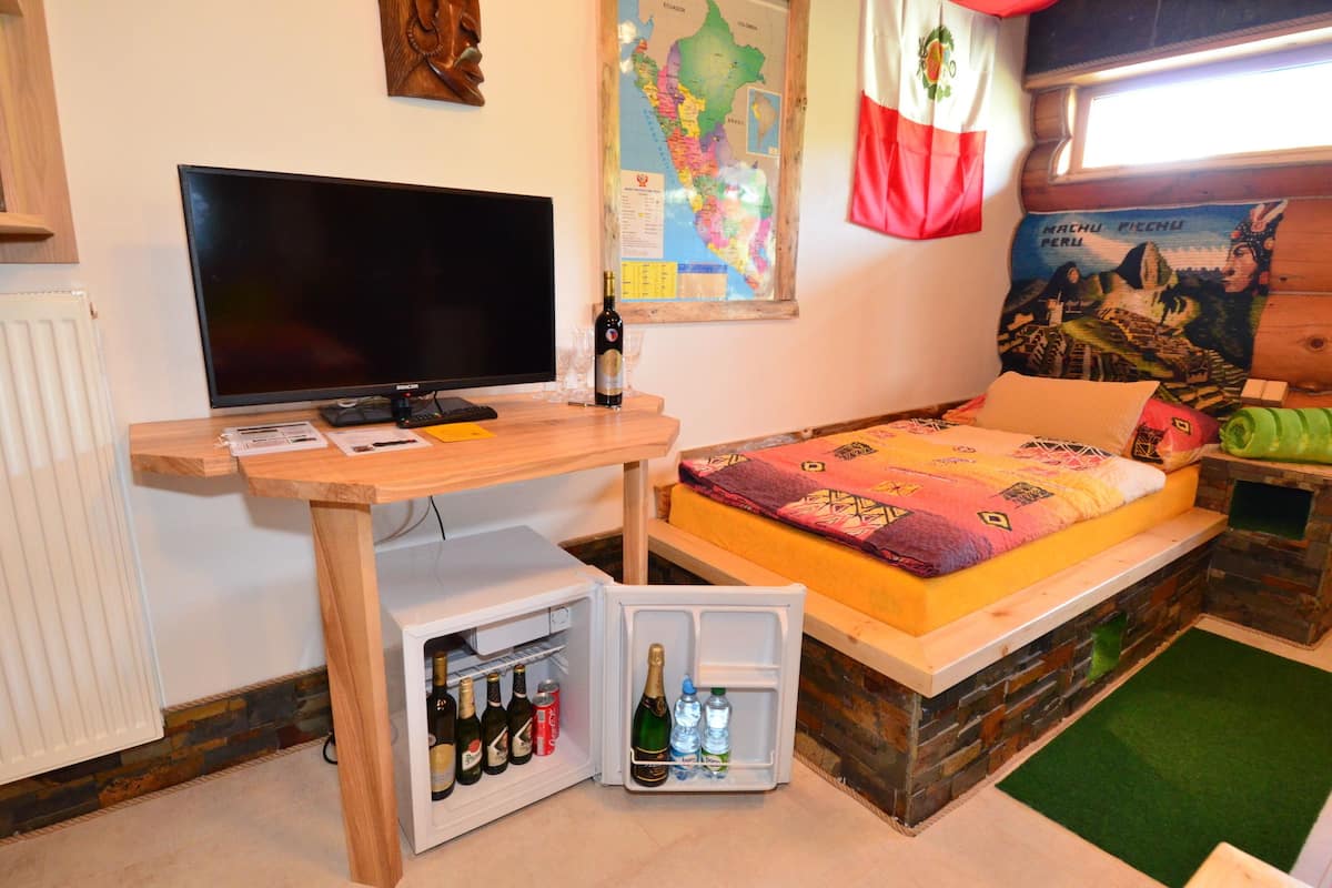 A compact room with a bed, a small wooden desk with a TV, a mini fridge stocked with beverages, a map on the wall, and a colorful bedspread.