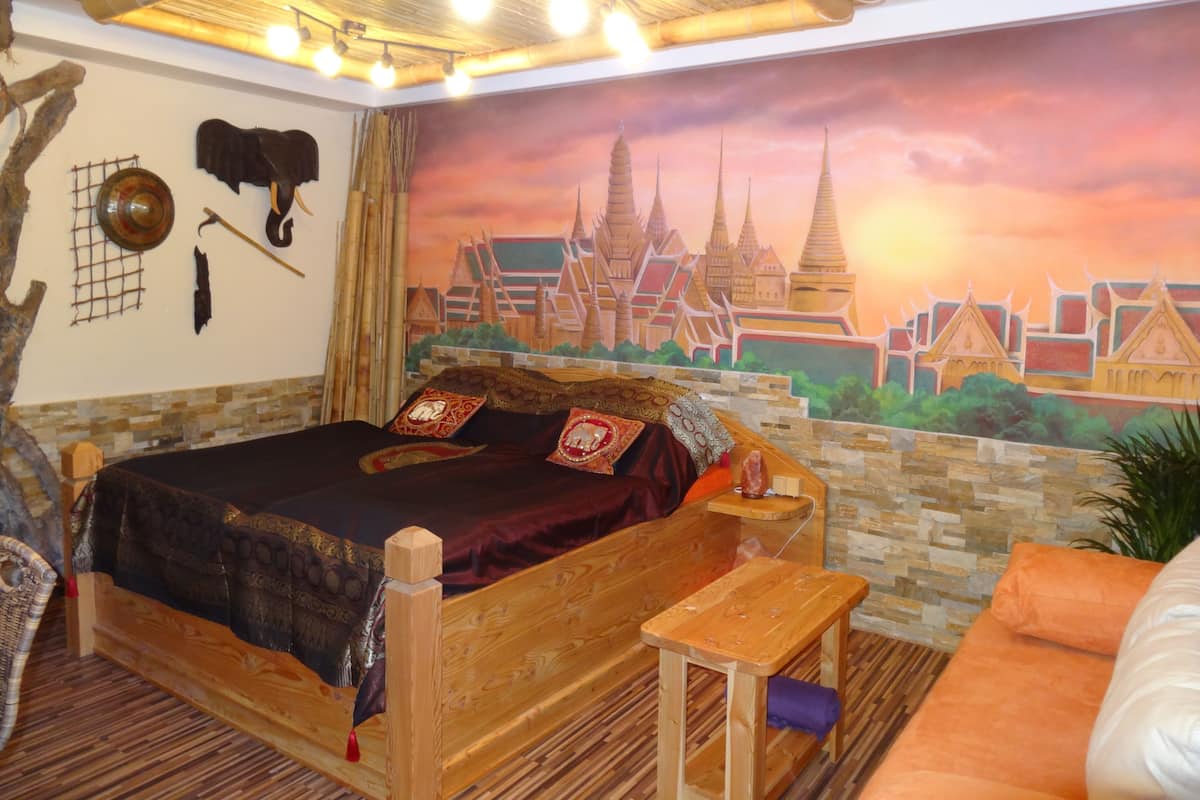 A room with a bed, a small wooden table, a bench, a wall with a mural, and a hanging elephant head.