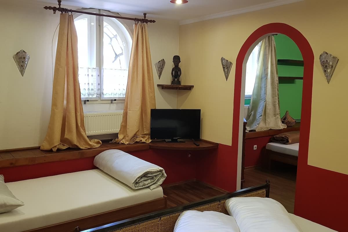 A room with a bed, a television, a window with curtains, a small shelf with a statue, and a mirror.