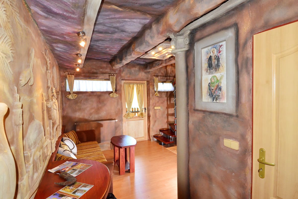 A room with a wooden ceiling, a bed with a striped blanket, a small stool, and a door with a light switch.
