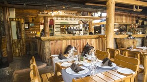 Un interior rústico de madera con mesas dispuestas para comer, una zona de bar con varias botellas y copas, y una chimenea de piedra.
