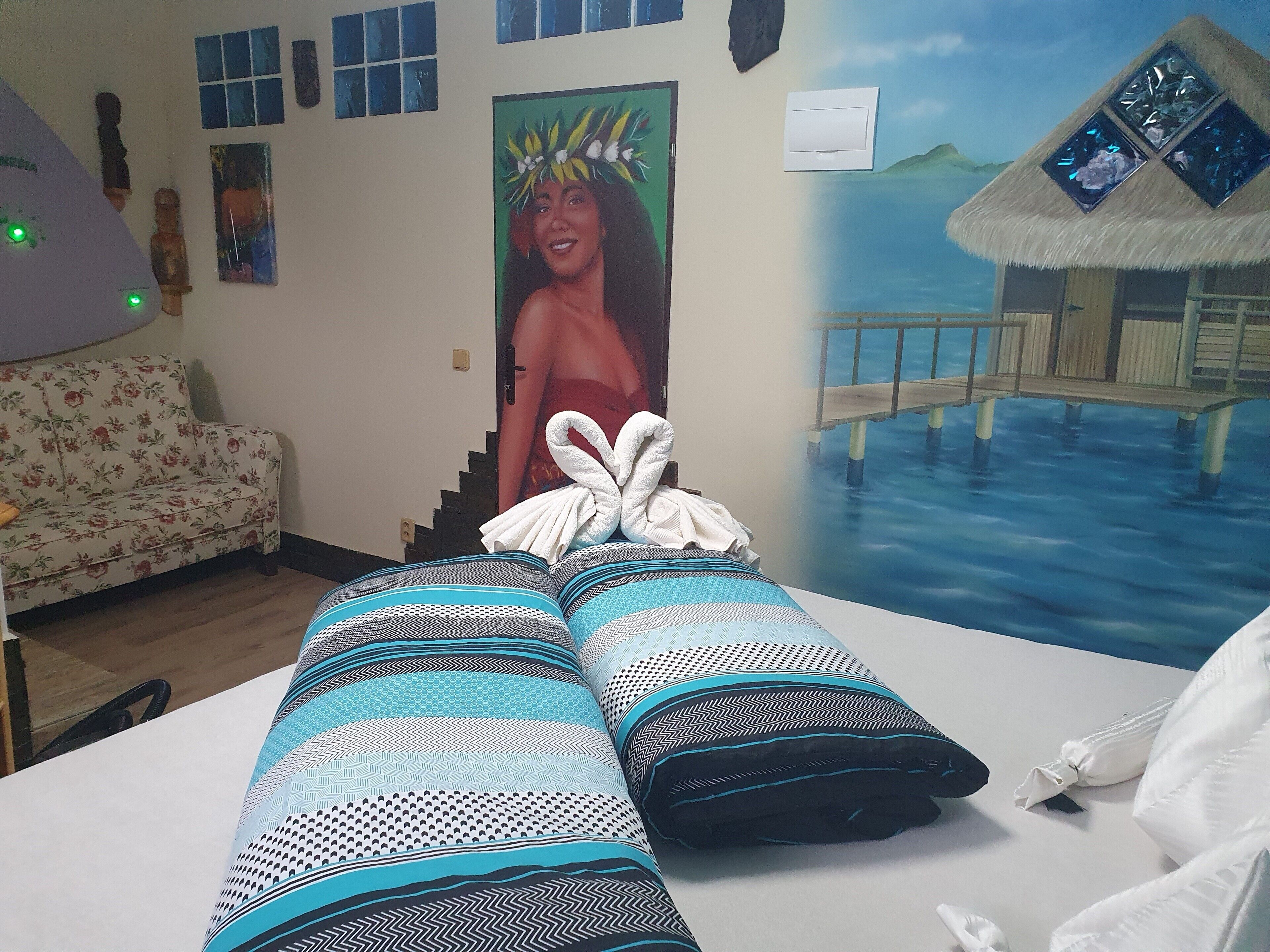 A room with a bed, a painting of a tropical scene, and a thatched-roof structure.