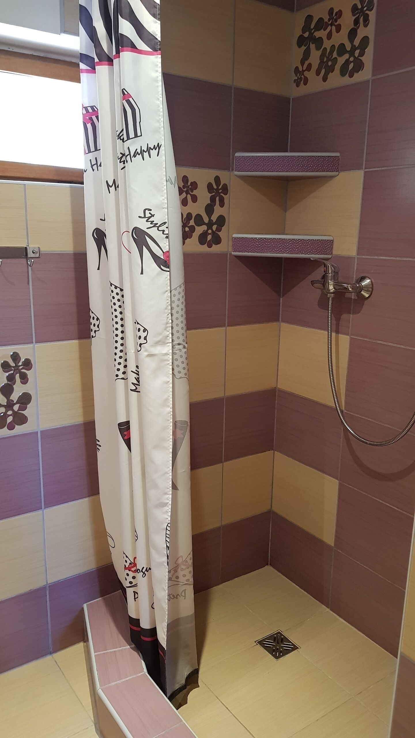 A shower area with a white curtain featuring various patterns and text, tiled walls with floral designs, and a showerhead.