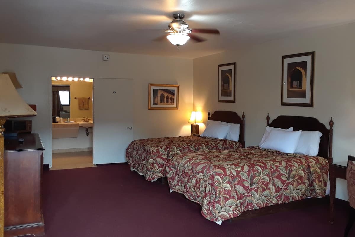 A hotel room with two beds, a ceiling fan, a nightstand with a lamp, a dresser, and a bathroom visible through an open door.