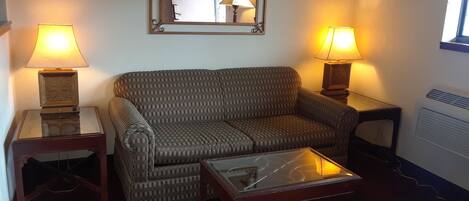 A living room with a patterned sofa, two side tables with lamps, a glass coffee table, and a mirror on the wall.