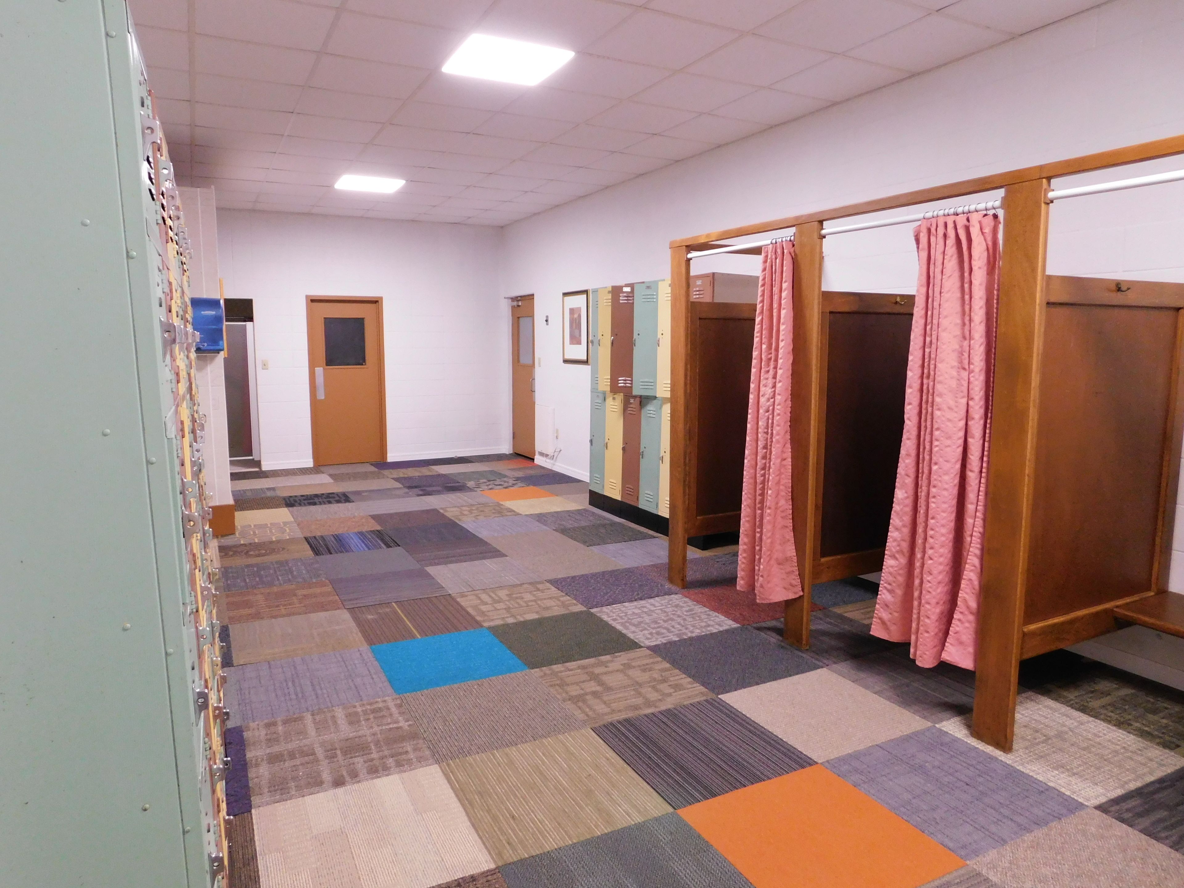 A hallway with lockers, a patterned carpet, and wooden cubicles with pink curtains.