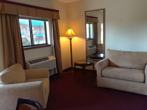 Habitación de hotel con un sofá beige, una mesita redonda pequeña, una lámpara de pie y un ventanal con cortinas.