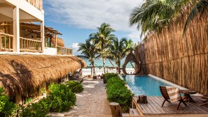 A pool area with a thatched-roof structure, palm trees, and a wooden deck with chairs. - Dune Boutique Hotel located at the party zone (Tulum)